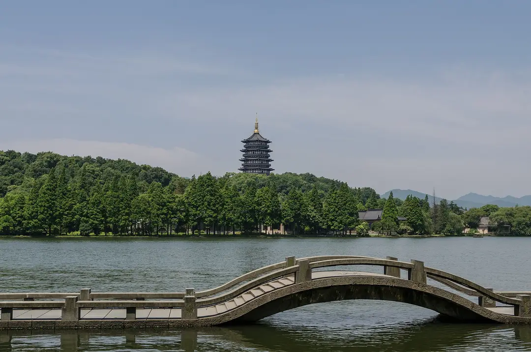 Leifeng Pagoda on a wooded hill above West Lake, with an arched stone footbridge in the foreground