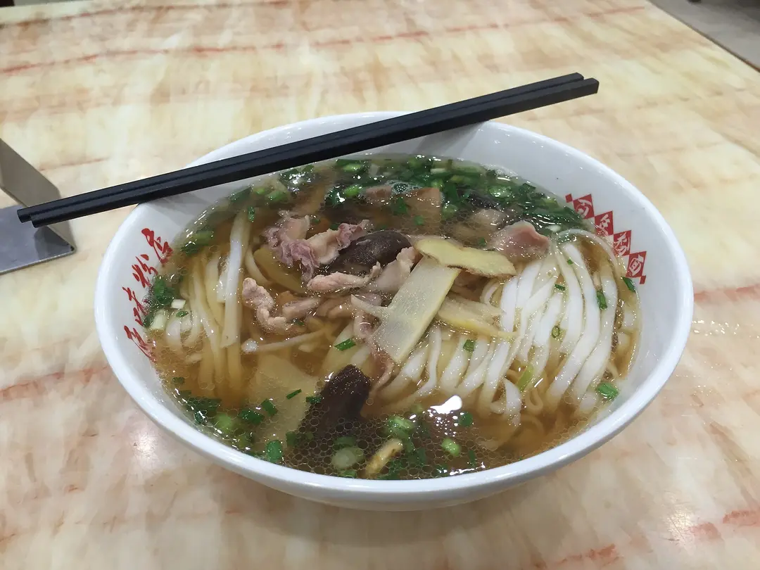 A bowl of flat rice noodles in clear broth with sliced meat and spring onion