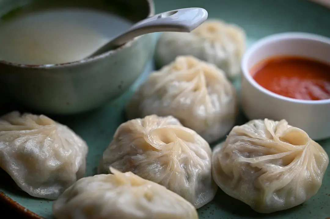 Pleated steamed momos on a green plate with a bowl of soup and a dish of red chili sauce