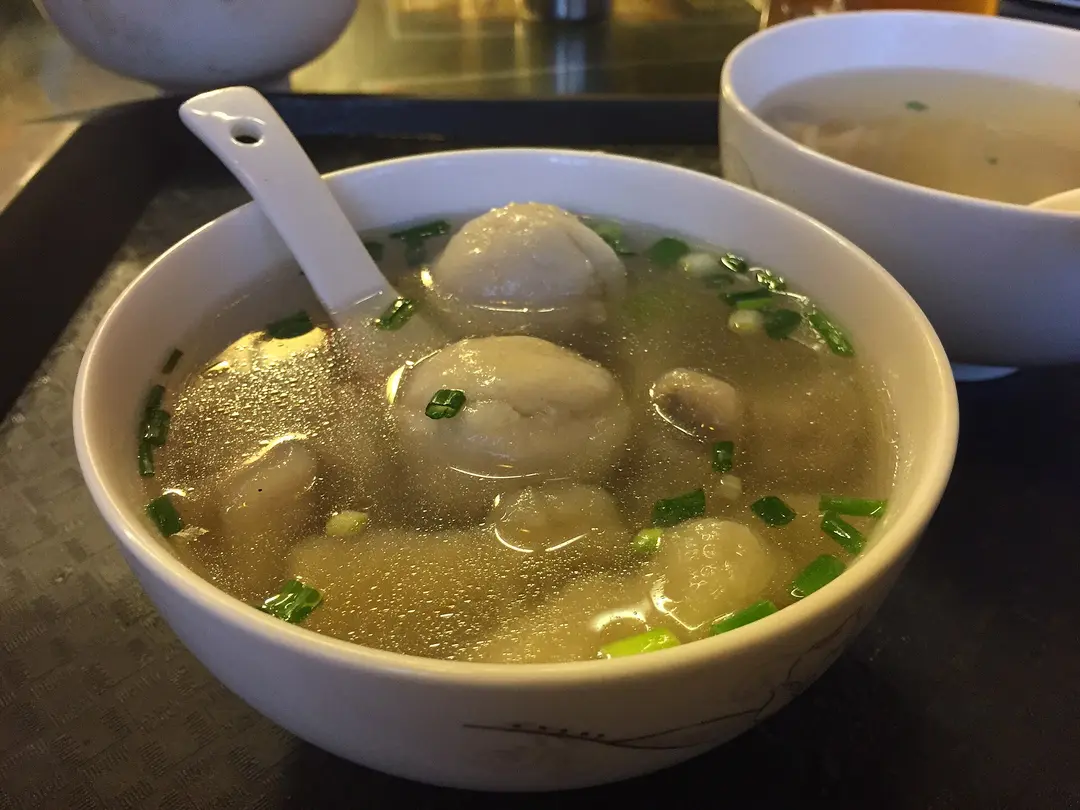 A bowl of Fuzhou fish balls in clear soup with chopped scallions and a white spoon