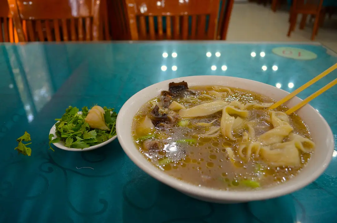 A bowl of wide wheat noodles in broth with meat, with a side dish of cilantro
