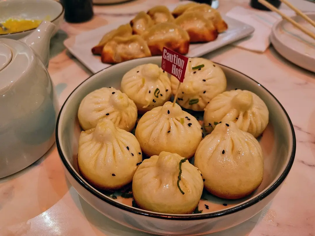 A bowl of pan-fried shengjian buns topped with sesame and scallion, with a teapot beside it