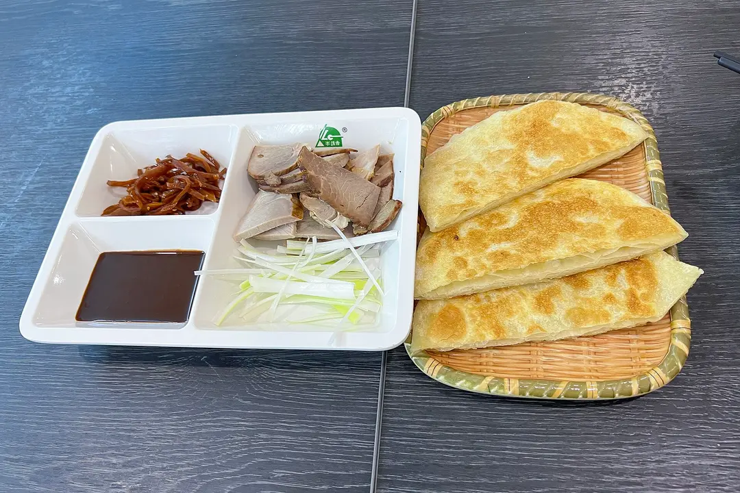 Tray of sliced smoked pork, scallion and sauce beside three wedges of golden pancake