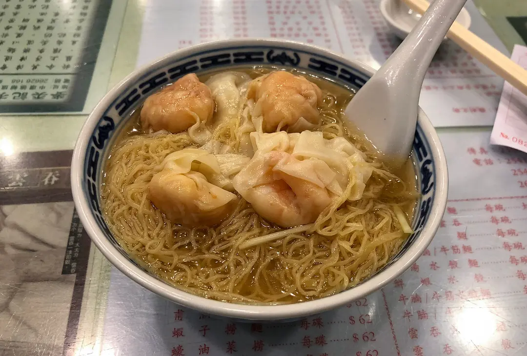 A bowl of shrimp wontons on thin egg noodles in clear broth, with a white spoon