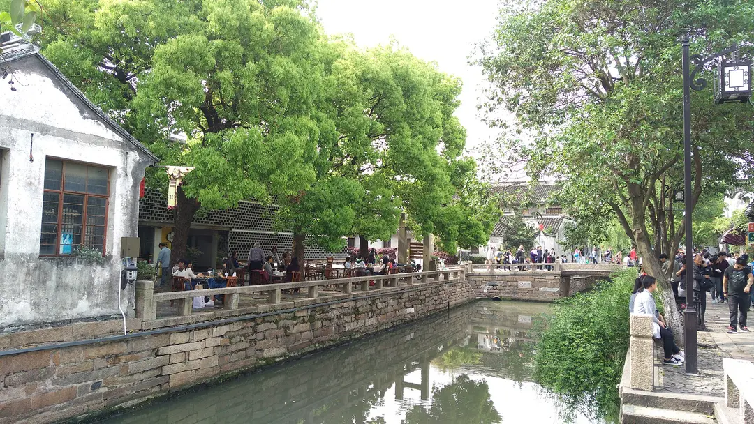 A stone-banked canal on Pingjiang Road, with café tables under camphor trees and a small bridge