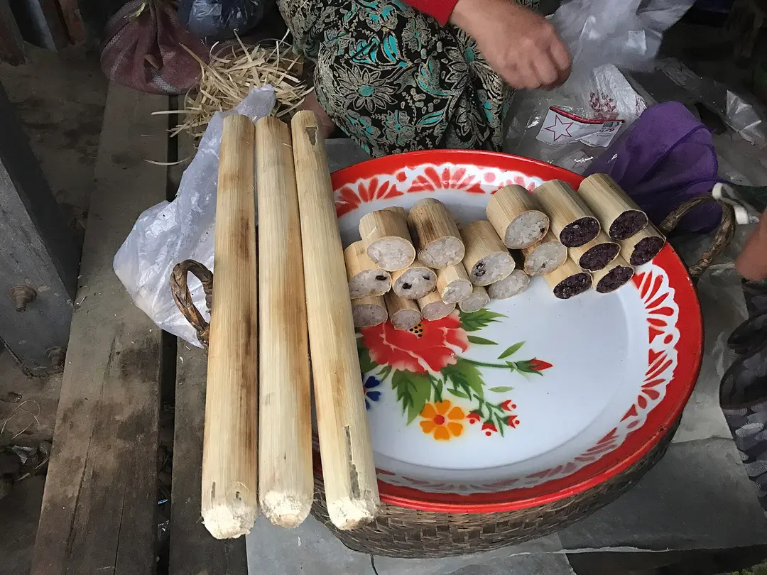 Whole bamboo tubes and cut pieces filled with white and purple sticky rice on a tray at a market