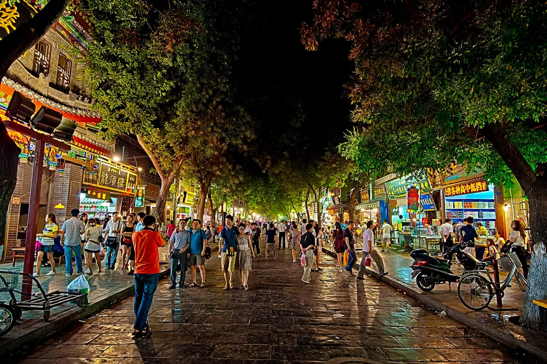 A tree-lined food street in the Muslim Quarter at night, lit by shop signs and crowded with people