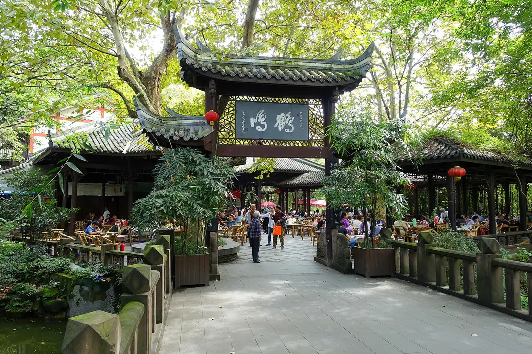 A paved path through the open-air teahouse in People's Park, Chengdu, under tall trees and a carved gateway