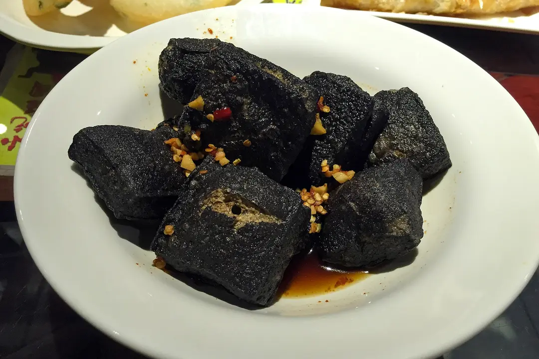 Black Changsha-style stinky tofu with chili sauce and chopped garlic on a white plate