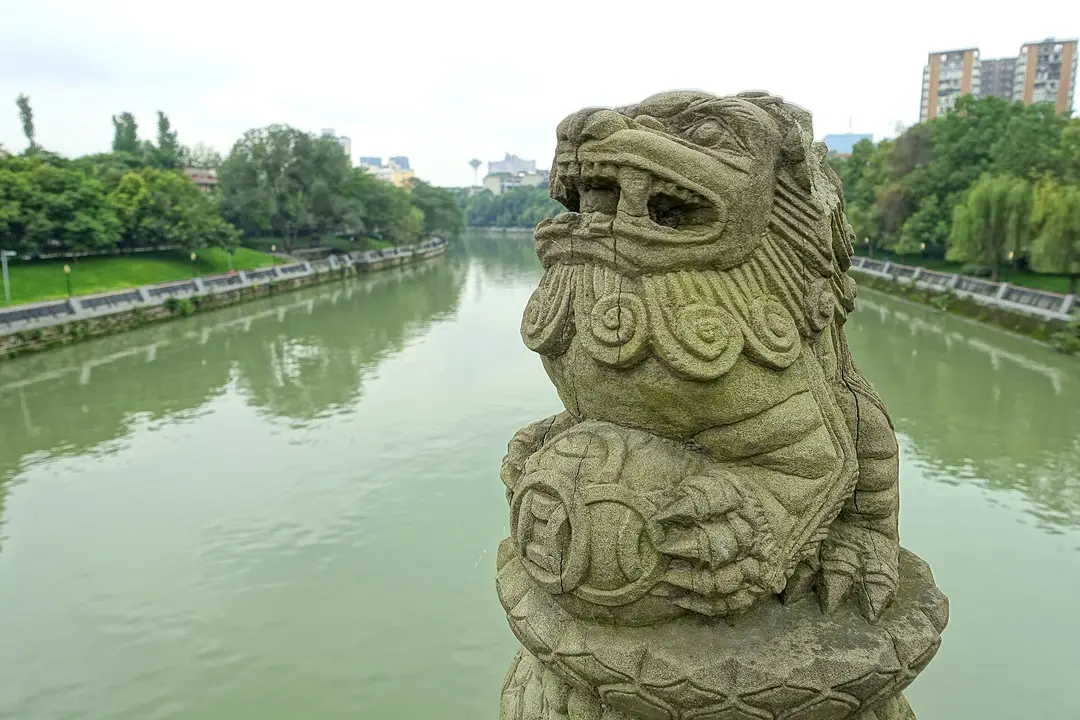 A carved stone lion on a bridge above the tree-lined Jinjiang River in Chengdu
