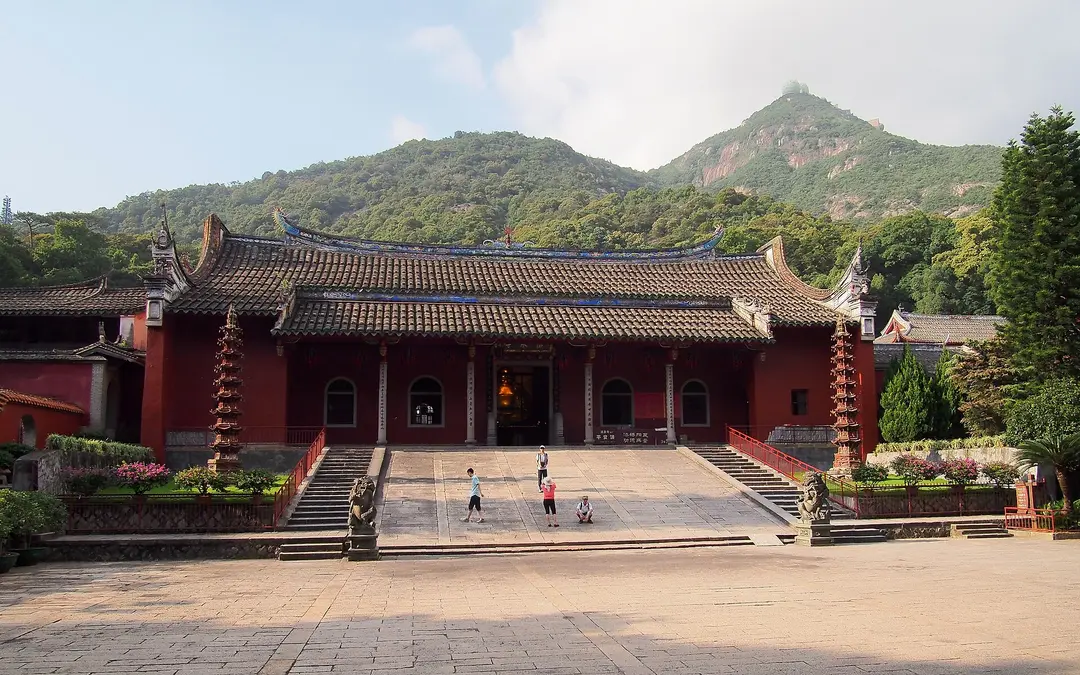 A red temple hall with curved roofs and two pagodas at Yongquan Temple, below the green peaks of Gushan