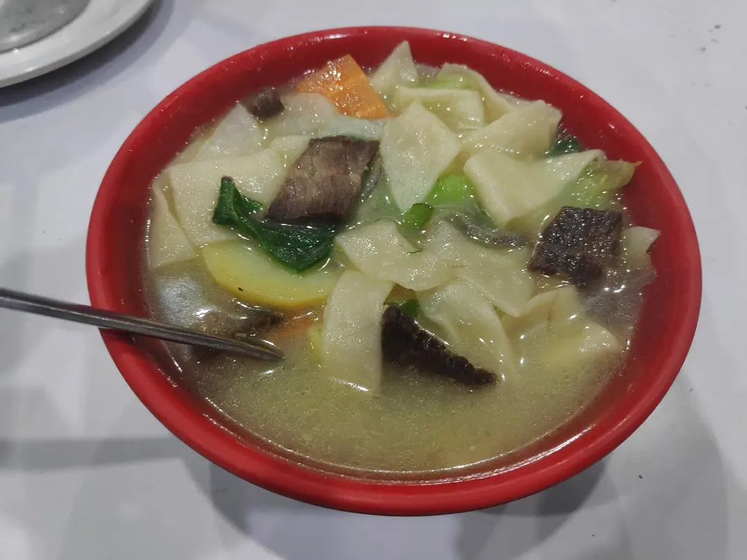 A red bowl of thenthuk with flat torn noodles, meat, potato and greens in clear broth