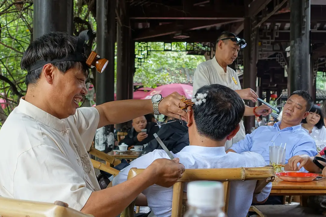 An ear cleaner with a head lamp working on a seated customer at a teahouse table in People's Park