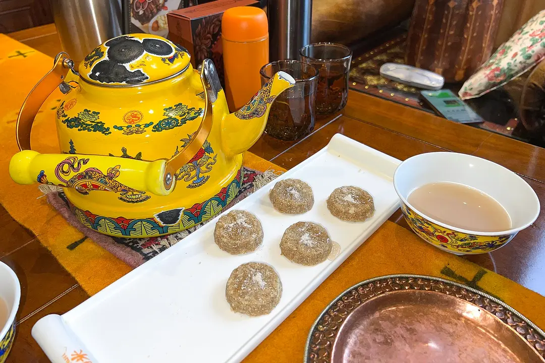 Five round tsampa cakes on a white tray beside a yellow Tibetan teapot and a bowl of butter tea