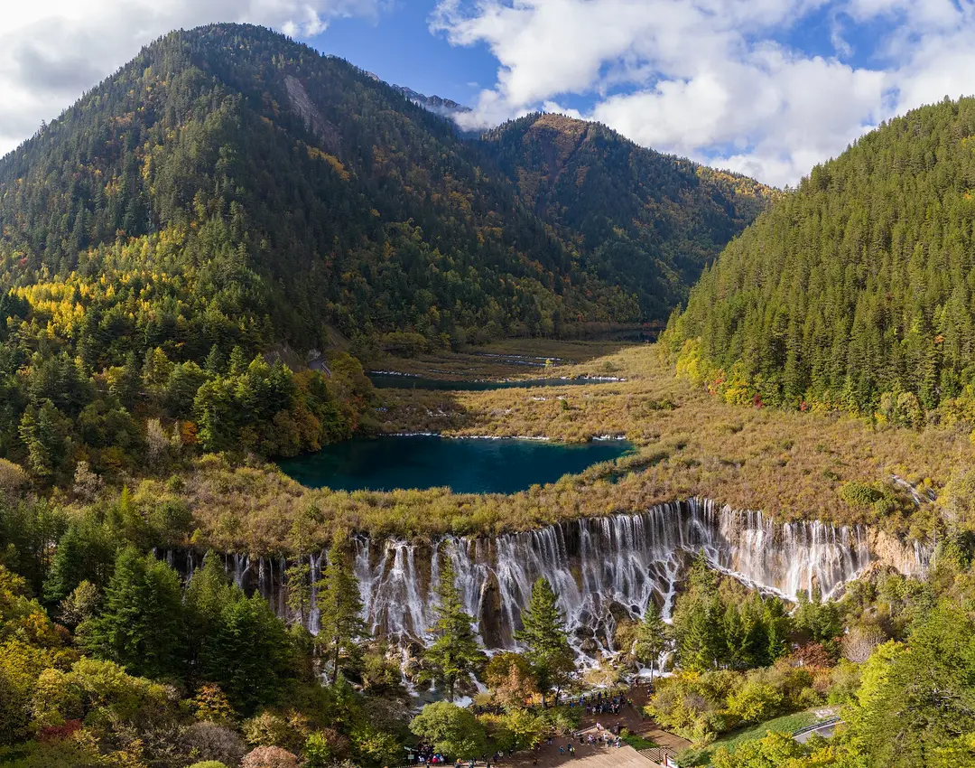 Nuorilang Falls spilling over a wide wooded ledge below a blue lake and forested mountains