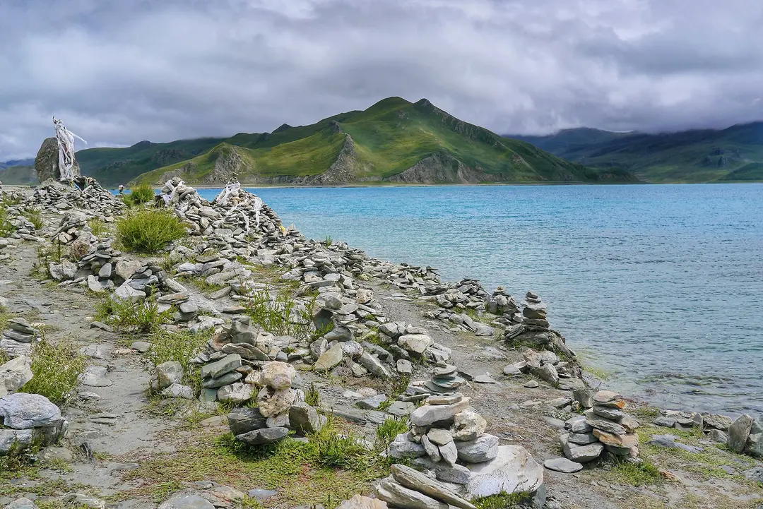 Stone cairns along the shore of turquoise Yamdrok Lake, with green hills under low cloud
