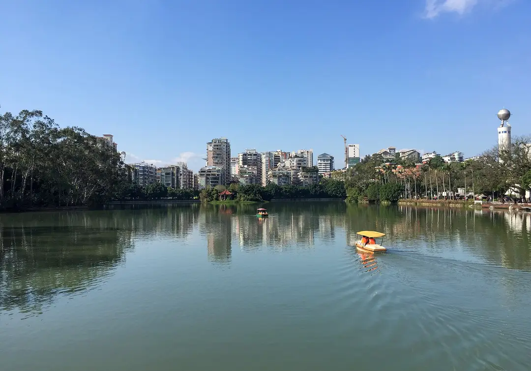 A small pedal boat crossing Fuzhou's West Lake under a blue sky, with trees and apartment towers on the far shore