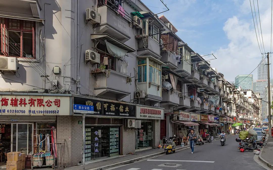 Small shops with Chinese signs under old apartment balconies on Xinyongan Road, Shanghai, with towers behind
