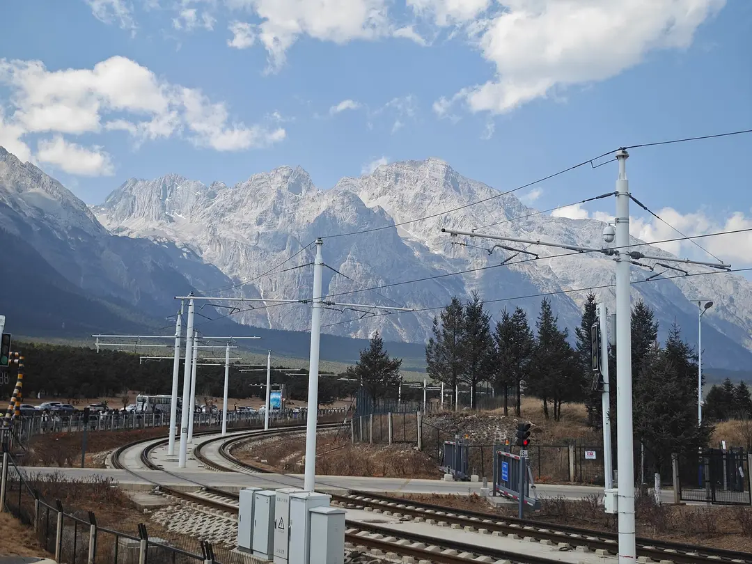 The snowy ridges of Jade Dragon Snow Mountain above rail tracks and pine trees on the plain below