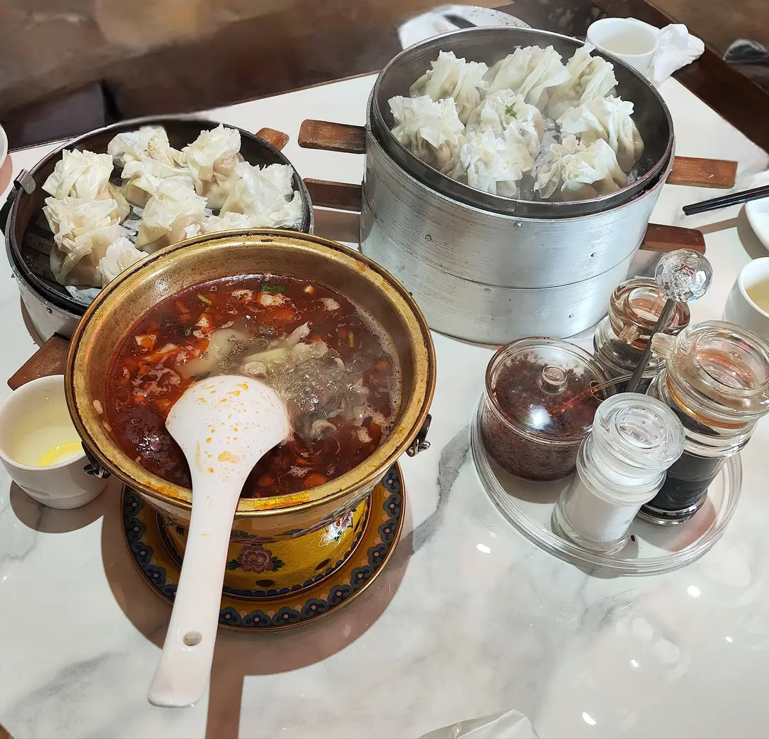 Two steamers of open-topped mutton shaomai beside a copper pot of red, chili-oiled lamb offal soup