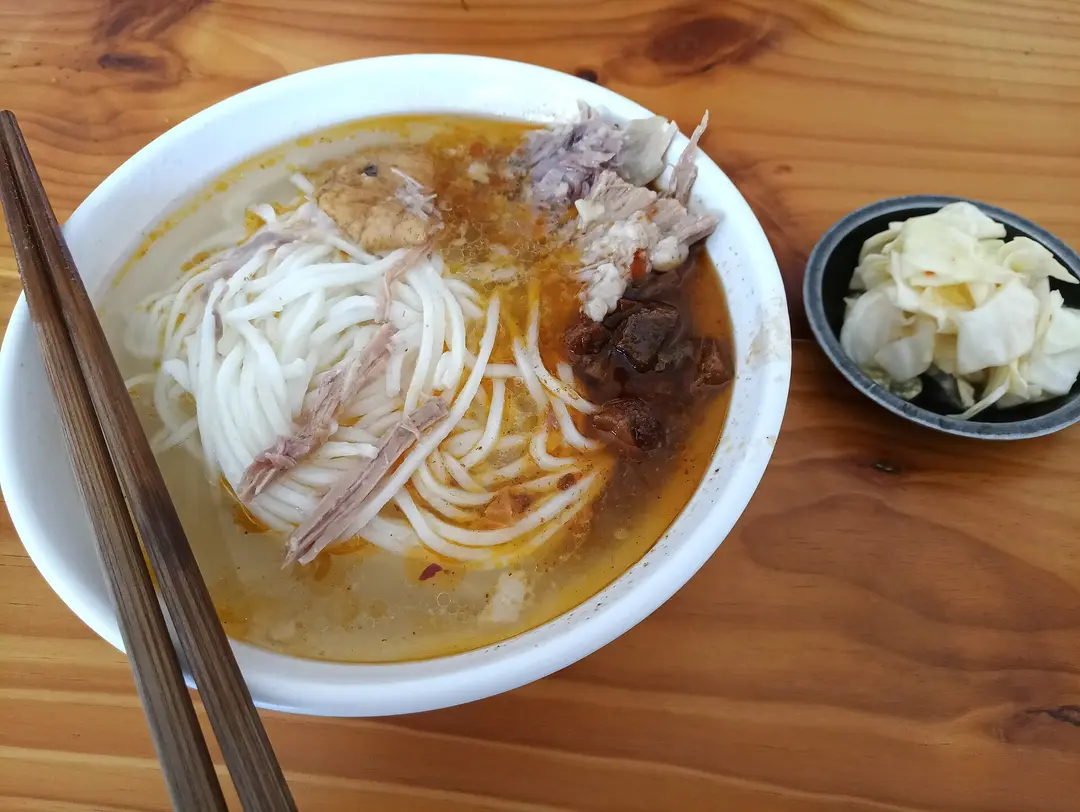 A bowl of rice noodles in broth topped with stewed pork, with pickles on the side