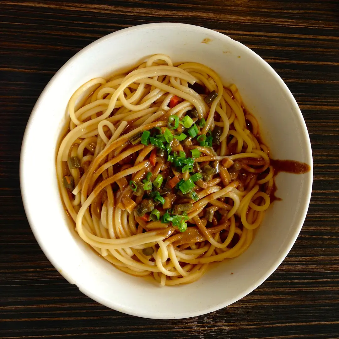 A bowl of hot dry noodles topped with sesame sauce, pickles and chopped scallions