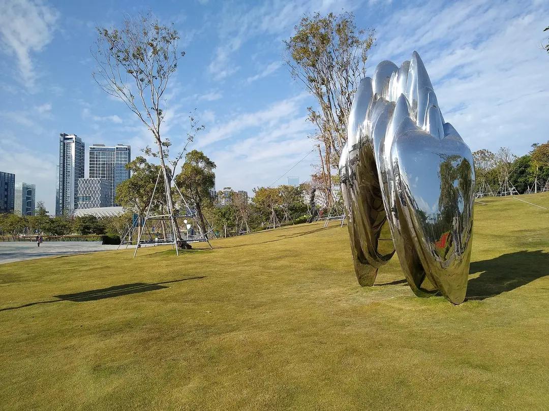A mirrored steel sculpture on the lawn at Shenzhen Bay Park, with office towers behind